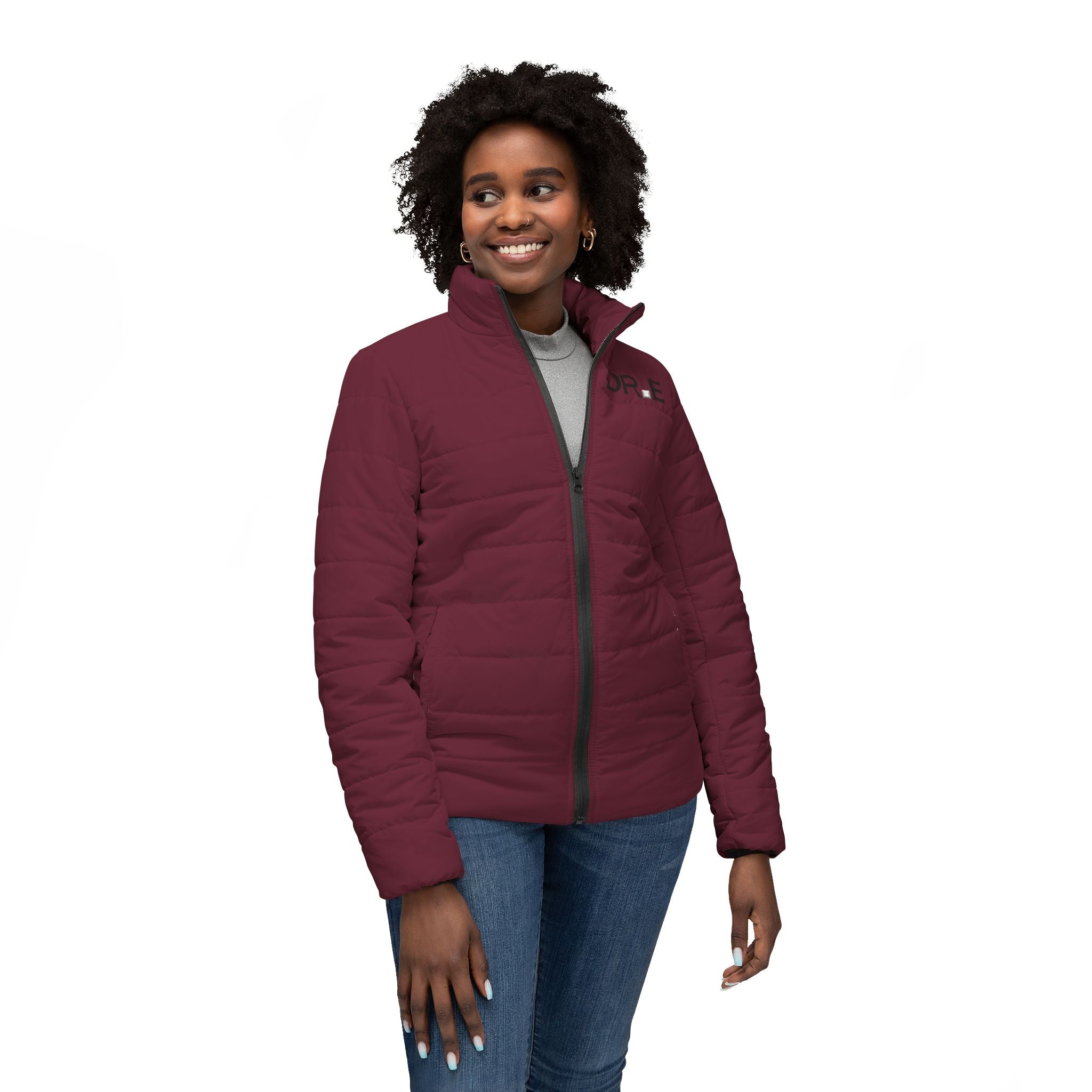 A woman wearing a burgundy quilted puffer jacket with a black zipper, styled with a gray top and blue jeans, photographed against a solid white background.